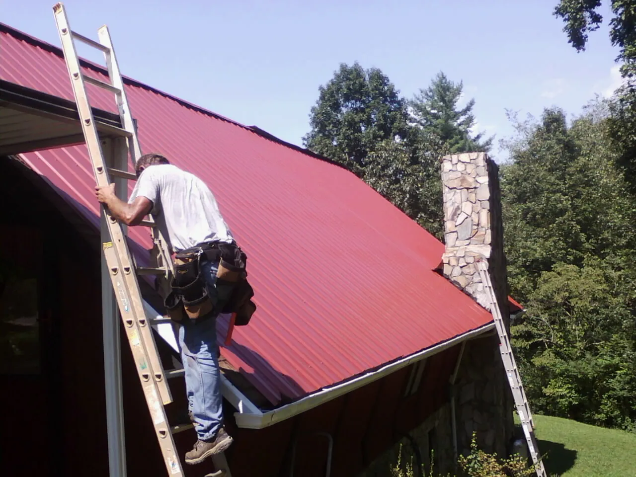 Roofer on a ladder beside a red metal roof and stone chimney — Soffit & Fascia Repair by Roofing Brothers in Parkersburg